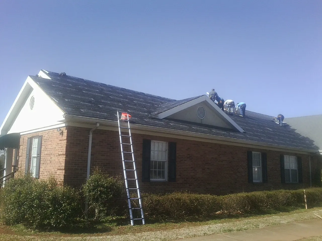 Crew tearing shingles off a brick ranch roof with a ladder set — Metal Roof Repair work by our crew in Eyota