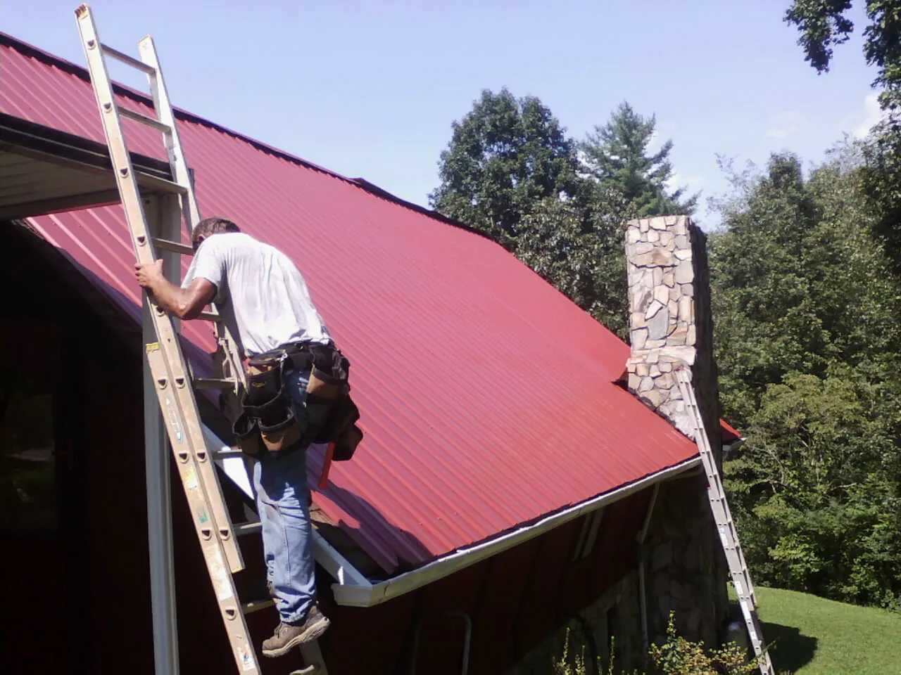 Roofer on a ladder beside a red metal roof and stone chimney — Soffit & Fascia Repair by Roofing Brothers in Eyota